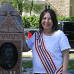 A woman in a Métis sash stands beside a Louis Riel monument.