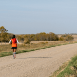 A woman in an orange vest runs away from the camera down a gravel road.