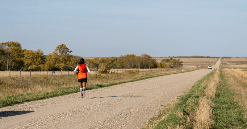 A woman in an orange vest runs away from the camera down a gravel road.