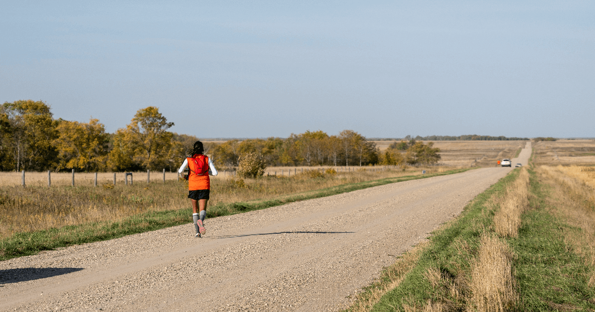 A woman in an orange vest runs away from the camera down a gravel road.