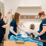 Nursing students working with a manikin in a hospital-style bed in a simulation lab.