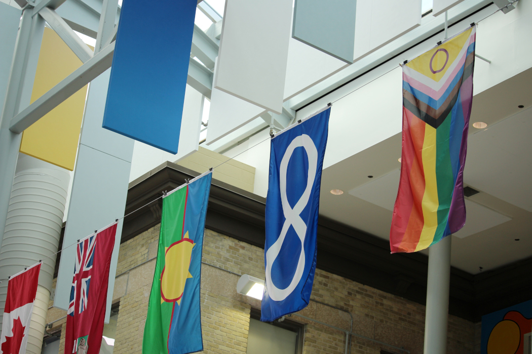 The Pride flag hangs from a wire in Brodie Centre atrium next to the Métis flag, Treaty One flag, flag of Manitoba, and flag of Canada.