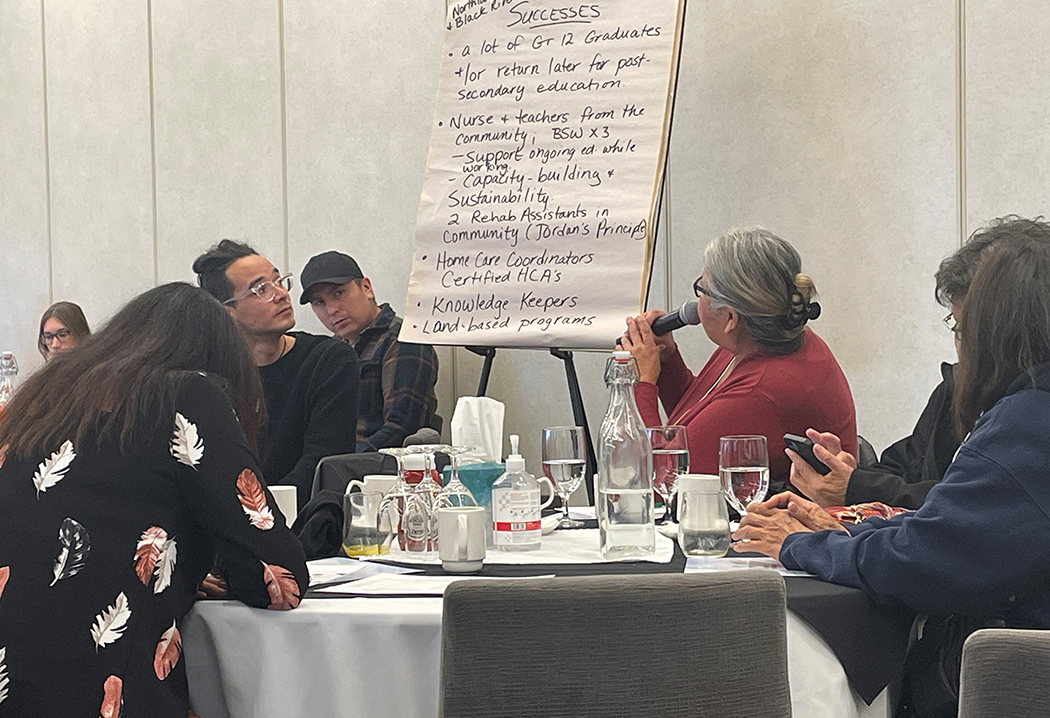 A group of people from First Nations communities have a discussion around a table with a list on a flip chart under the heading "successes."