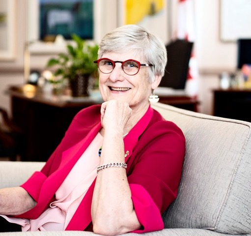 Senator Patricia Bovey in wearing a pink/red sweater siting in a chair smiling toward the camera.