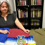 An Indigenous woman sits behind a table of colourful ribbon skirts.