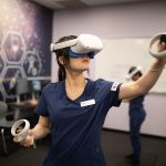 A nursing student wears a virtual reality headset and holds virtual reality hand controllers.