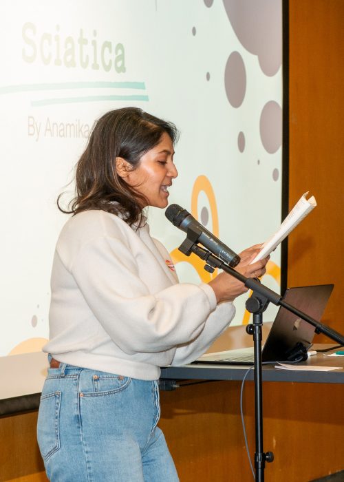 Female standing at a microphone reading from a book in her hands.