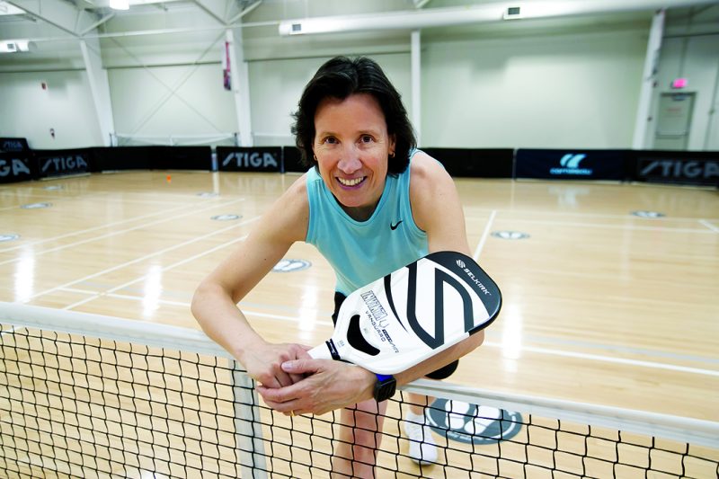 Sandra Webber leans over a pickleball net, holding a racquet.