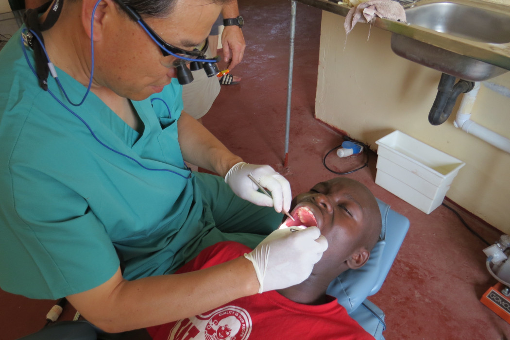 Patient in a dental chair with their mouth open. Dr. Kim performs dental treatment while wearing white gloves and holding dental instruments.