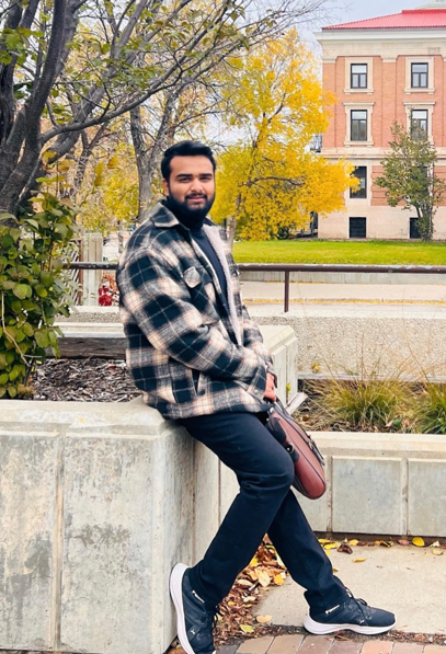 Man leaning on stone wall, holding a leather satchel. University of Manitoba administrative building in the background.