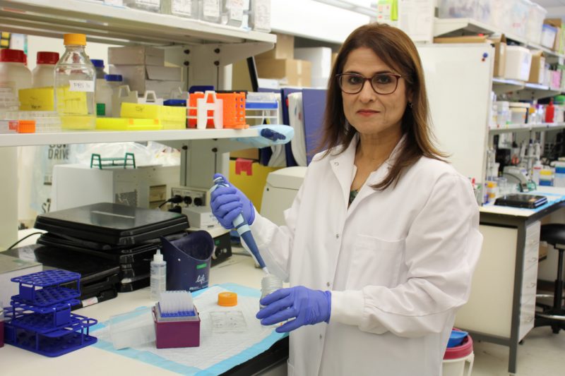 Dr. Soheila Karimi holds a pipette and a jar. She is in her lab and is wearing gloves and a lab coat.