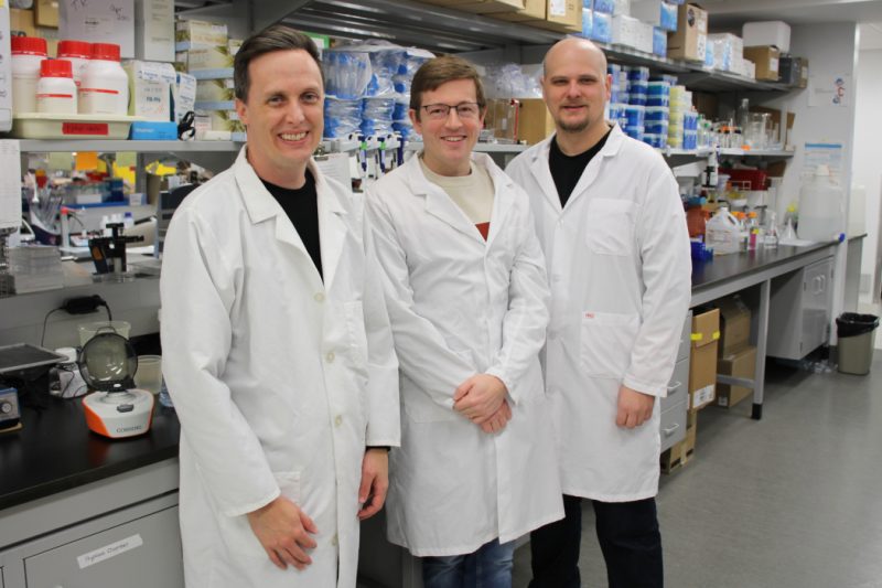 The three researchers pose in a lab. They are wearing white lab coats.