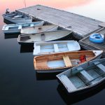 Seven two-man fishing boats, of a variety of sizes, styles, materials, and ages sit tied up to a weathered wood dock on a perfectly calm lake during sunset.