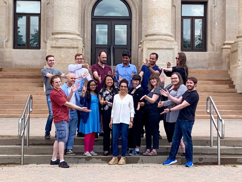 Dr. Samar Safi Harb standing in the middle of her research group smiling with students around her pointing to her in front of the UM Administration building.