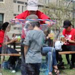 A kid with a grey t-shirt and cap standing in the middle of a large bubble created around him by a student wearing a red shirt with soap and hula loop in front of the science buildings complex outside on the grass with kids and other people in the background.