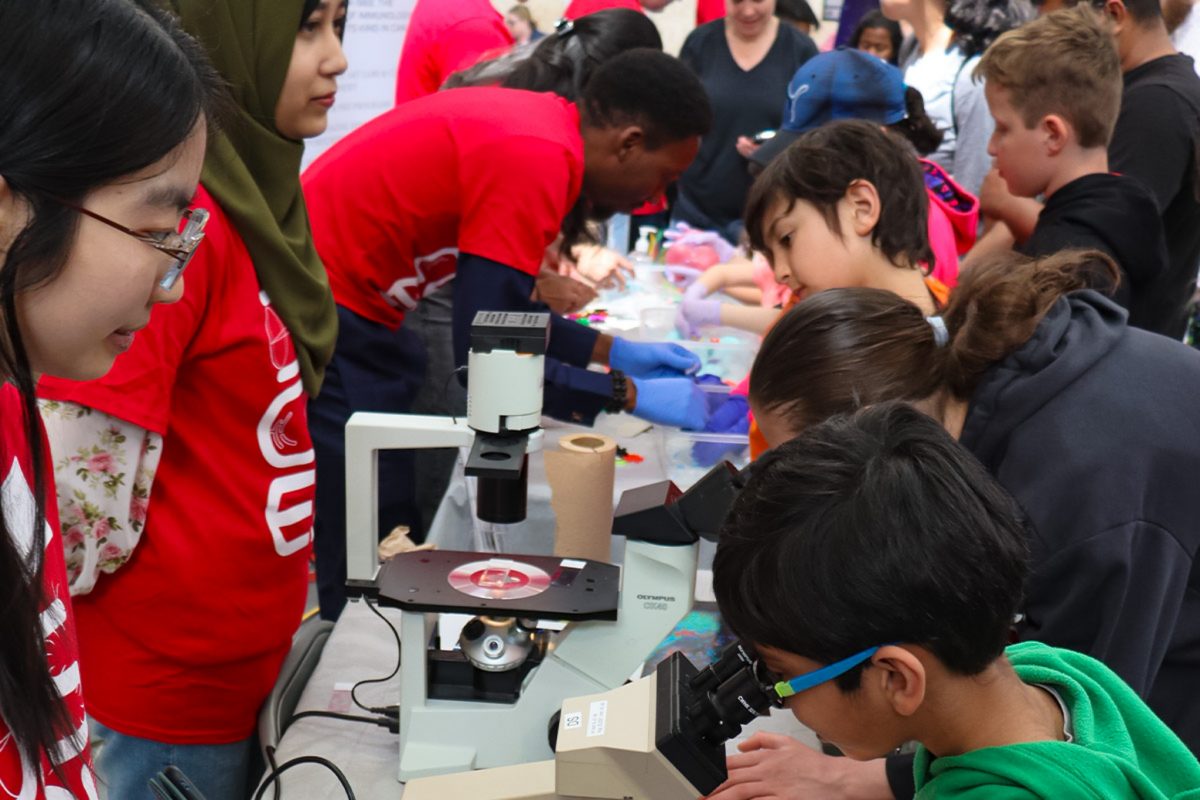 University students and kids on both sides of a table with microscopes while some kids are looking through the microscopes and some university students are explaining or showing the science experiments to kids.