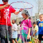A kid in the middle of a bubble made with soap and hula loop by a university student wearing a red Science Rendezvous volunteer shirt.
