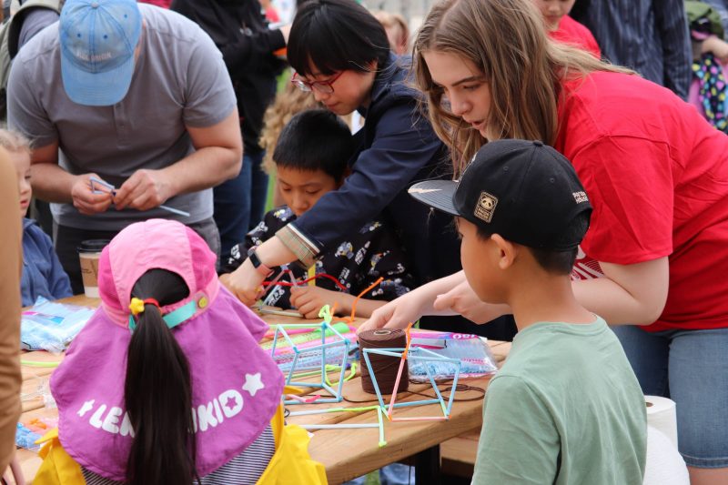 A university students with long hair and a red shirt working with colorful plastic straws to build structures and cubes while explaining it to the kids that are gathered around the table.