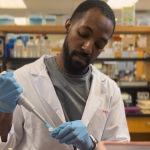 Steve Daley, Black PhD student, wearing a white lab coat with blue gloves on and doing experiments in the lab.