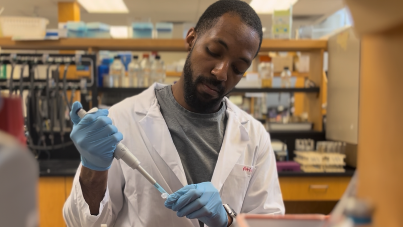 Steve Daley, Black PhD student, wearing a white lab coat with blue gloves on and doing experiments in the lab.