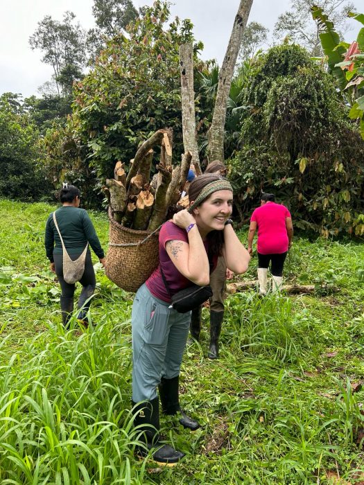 A young woman carries a basket of branches on her back in a rainforest setting.