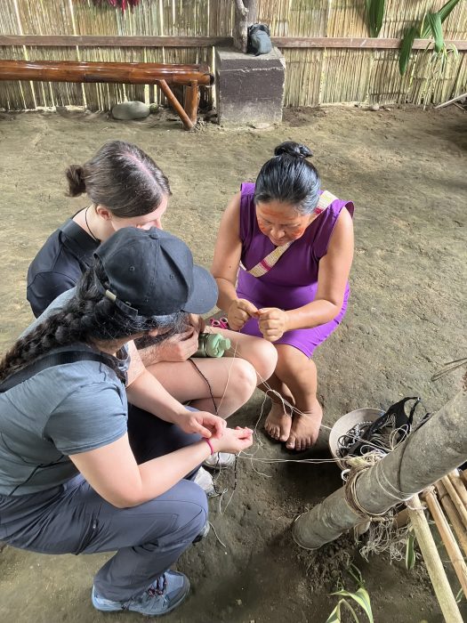 An Indigenous woman crouched down with two female university students, working with string.