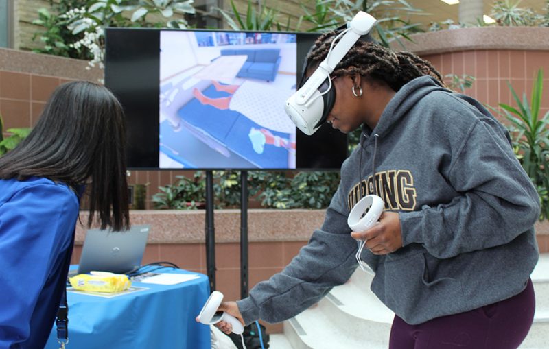 A woman wearing a VR headset and hand controls in front of a TV showing a virtual hospital room.