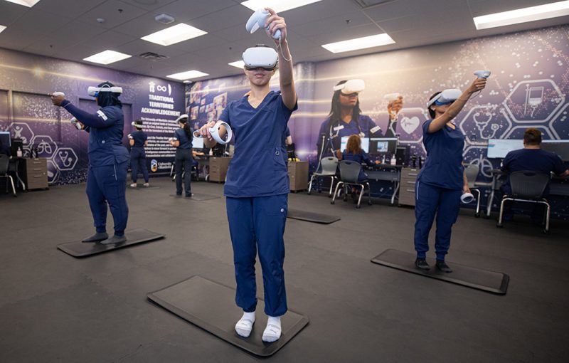 Nursing students in a lab setting wearing scrubs and virtual reality headsets and hand controls reach as they work in virtual environments. Other students in the background look at computer screens.