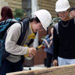 A student wearing a hardhat hammers a nail into a board as another student looks on.