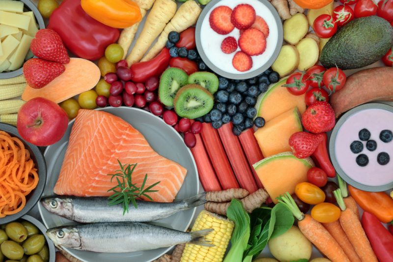 Healthy foods, including carrots, strawberries, red pepper, kiwi, blue berries, tomatoes, melon, salmon, avocado, olives and corn.