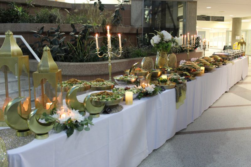 Food in bowls and buffet containers on long tables with tablecloths. Flowers and candles are also on the tables.