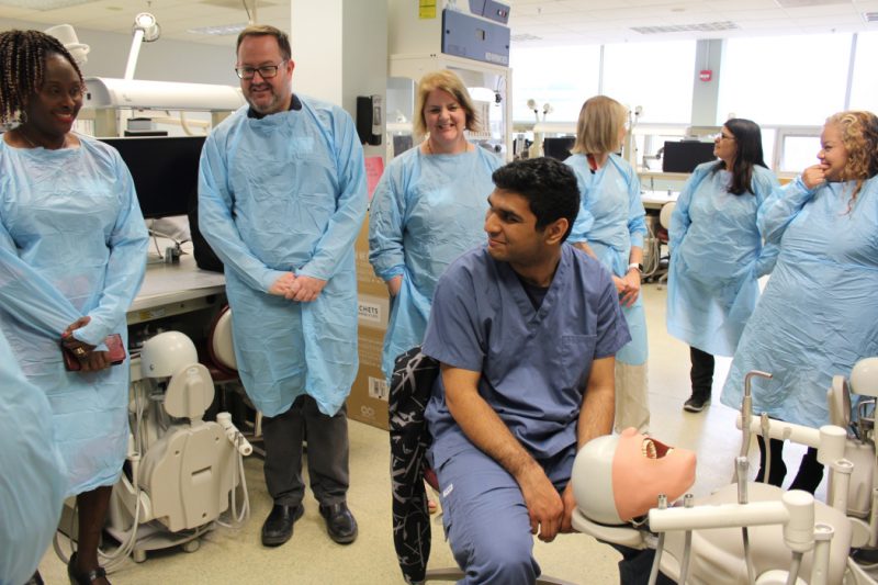 Six people wearing medical gowns stand around a student seated in front of a dental manikin. 