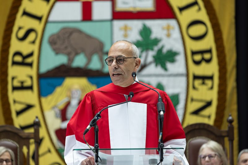 Graham Sher speaks at convocation in front of a large University of Manitoba banner.