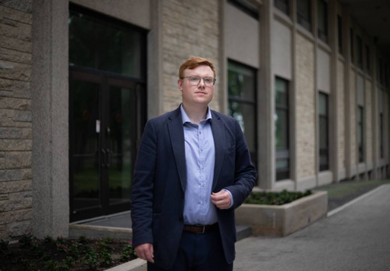 A man with red hair and glasses, wearing a light blue button down shirt and dark blue suit. He is standing in front of a building.