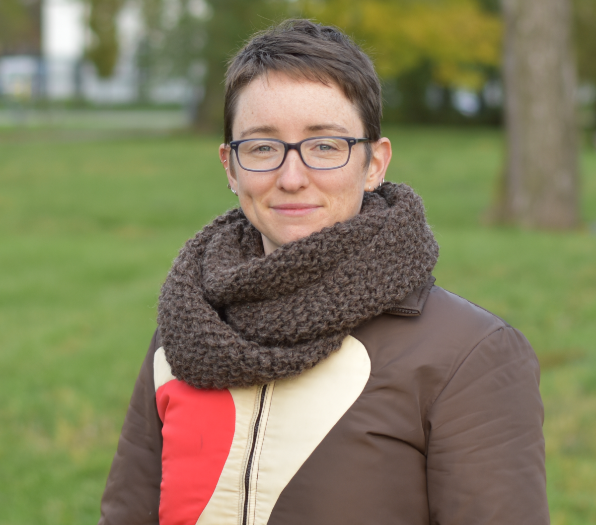 Woman with short dark brown hair, black glasses with a dark brown infinity scarf on, wearing a dark brown jacket with cream and red accents. She is standing outside in a park.