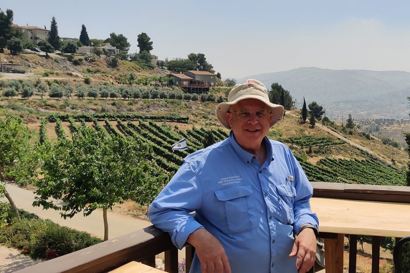 haskel greenfield, a man wearing a blue button up shirt and a beige hat, pictured on a wood deck with scenic hills behind in the distance.
