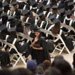 A UM graduate smiles and waves at the crowd during a convocation ceremony. Rows of grads in black caps and gowns face the stage behind her.