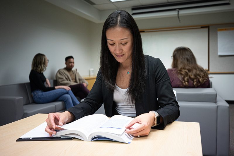 An Asian female student studies in a student lounge, with other students meeting behind her.