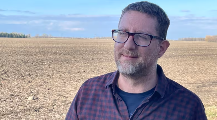 Man with short salt and pepper hair and beard, wearing black rectangular frame glasses. He is wearing a purple button down shirt with a dark blow shirt underneath. He is standing in front of a farmers field.