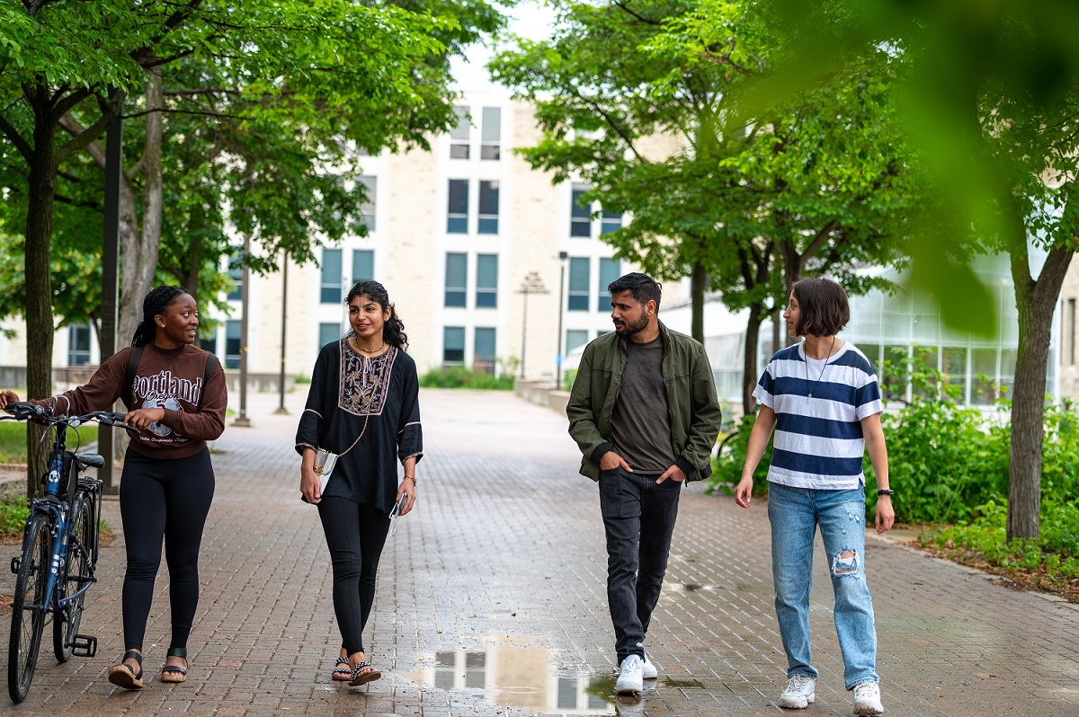 Four students walk along a campus pathway, one pushing a bicycle.