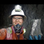 Woman in a white safety helmet with safety goggles, a breathing mask and orange protective clothing. She is standing in a mine holding a large chunk of stone.