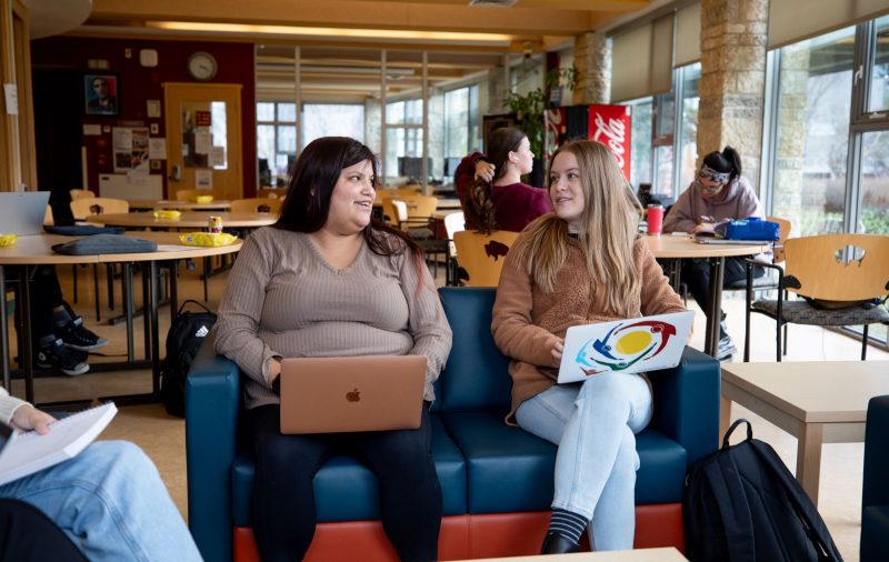 Two students with laptops sit on a couch in a student lounge. They are smiling at each other.