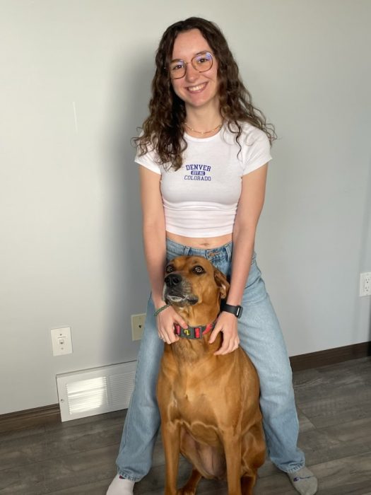 A student in a white t-shirt and jeans stands with her dog. She is smiling.