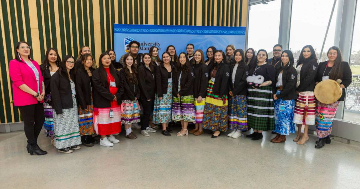 A group of students, many in ribbon skirts, stand together in a semi circle.