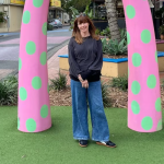 woman with should length brown hair, brown bangs. Wearing a long sleeve black sweatshirt, blue jeans and black sneakers. standing outside under a pink with green polka dots arch.