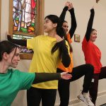 Four young women wearing different-coloured t-shirts perform movement exercises inside a church.
