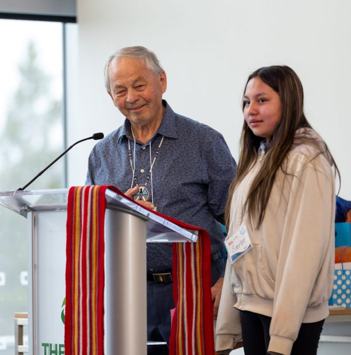 An Elder and his grand-daughter stand together at a podium that is adorned with a Métis sash.