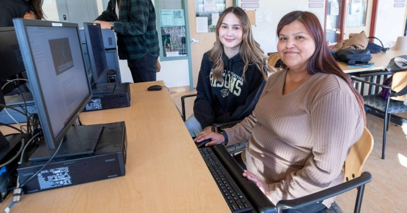 Two female students sit together at a computer, smiling at the camera.