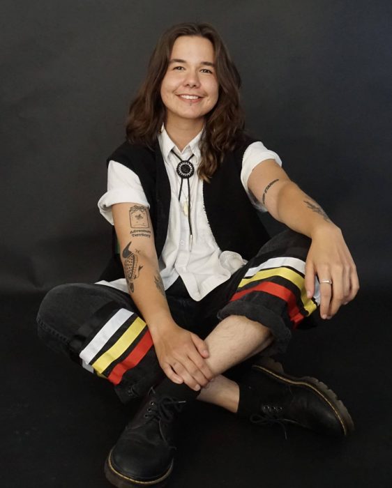 A student in sits cross-legged on a black background, facing the camera and smiling.
