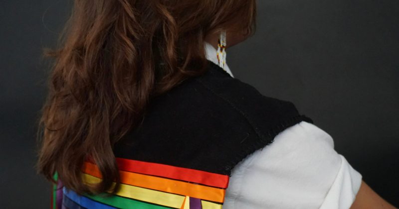 An Indigenous student with long hair poses with their back to the camera, only shoulders visible, wearing a black ribbon vest with a white t-shirt.